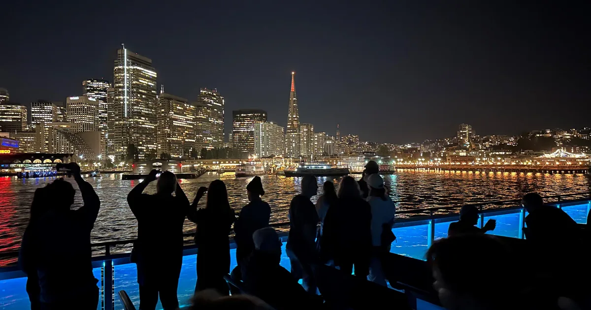 Passengers on deck photographing the San Francisco skyline lit up at night from a cruise on the bay