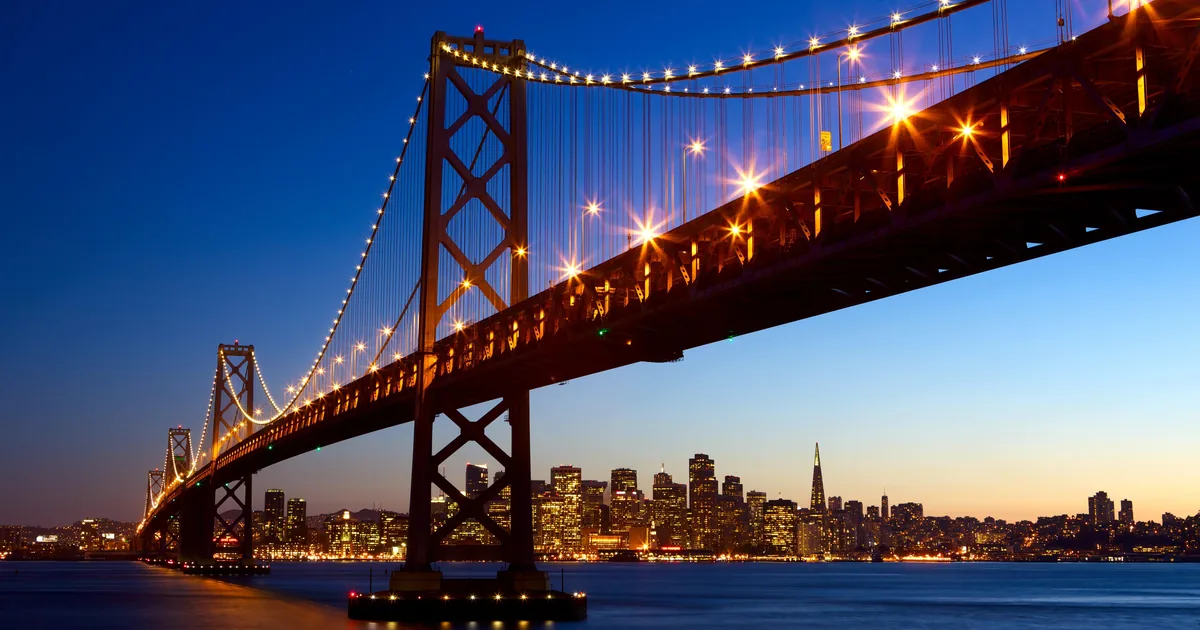 The Bay Bridge lit up at twilight with the San Francisco skyline and the Transamerica Pyramid beyond the water